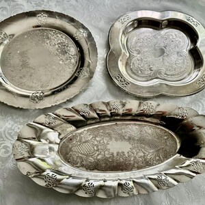 May include: Three silver serving trays with ornate designs. The trays vary in shape: a round plate with floral accents, a quatrefoil tray, and an oval tray with a scalloped edge. All have detailed embossed patterns.