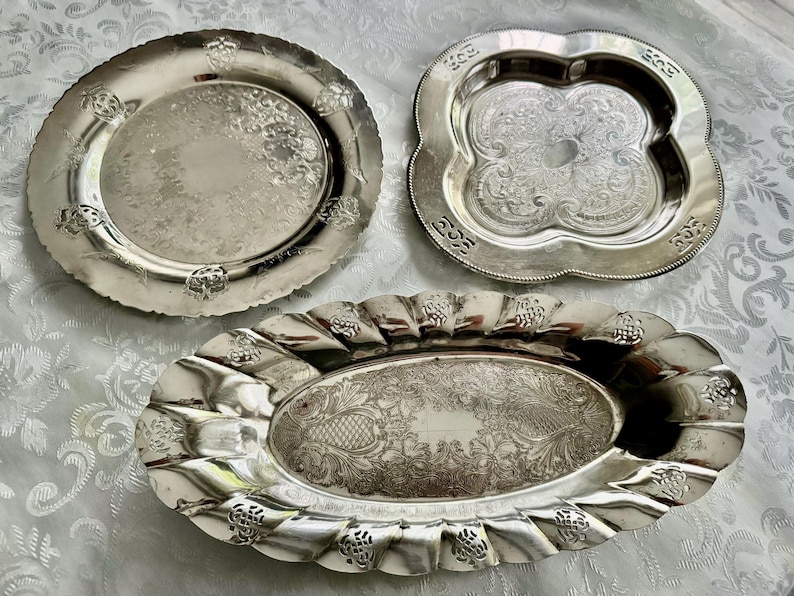 May include: Three silver serving trays with ornate designs. The trays vary in shape: a round plate with floral accents, a quatrefoil tray, and an oval tray with a scalloped edge. All have detailed embossed patterns.