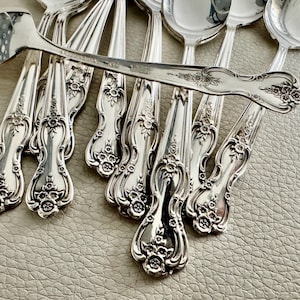May include: A collection of ornate silver spoons with floral detailing. The spoons are arranged on a textured surface, with one spoon prominently displayed on top. The spoons have a shiny, reflective surface.
