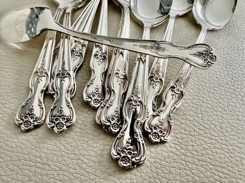 May include: A collection of ornate silver spoons with floral detailing. The spoons are arranged on a textured surface, with one spoon prominently displayed on top. The spoons have a shiny, reflective surface.