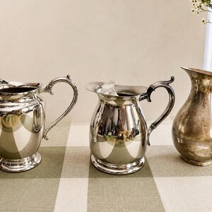 May include: Three silver-toned metal pitchers arranged in a row. The two pitchers on the left feature ornate, curved handles and wide pouring spouts, while the pitcher on the right has a simpler, tapered silhouette with a smooth, rounded body.
