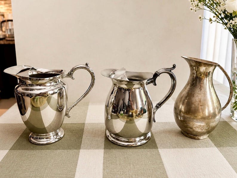 May include: Three silver-toned metal pitchers arranged in a row. The two pitchers on the left feature ornate, curved handles and wide pouring spouts, while the pitcher on the right has a simpler, tapered silhouette with a smooth, rounded body.