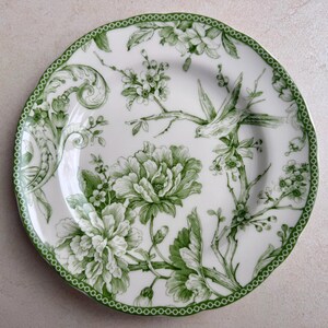 May include: A round ceramic plate featuring a white base with a green botanical pattern. The design includes detailed illustrations of blooming flowers, leafy branches, and a bird in flight, finished with a scalloped edge and a dotted border.