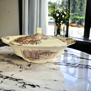 May include: A vintage cream-coloured ceramic serving bowl with a lid. The bowl features a brown and yellow floral pattern with a countryside scene.