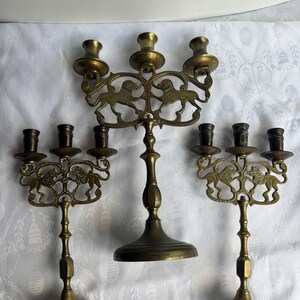 Pu&ograve; includere: Tre candelabri in ottone. Ogni supporto presenta un motivo decorativo a leone e pi&ugrave; portacandele. I candelabri sono di diverse altezze e hanno un'estetica vintage. I candelabri sono su una superficie bianca a motivi.