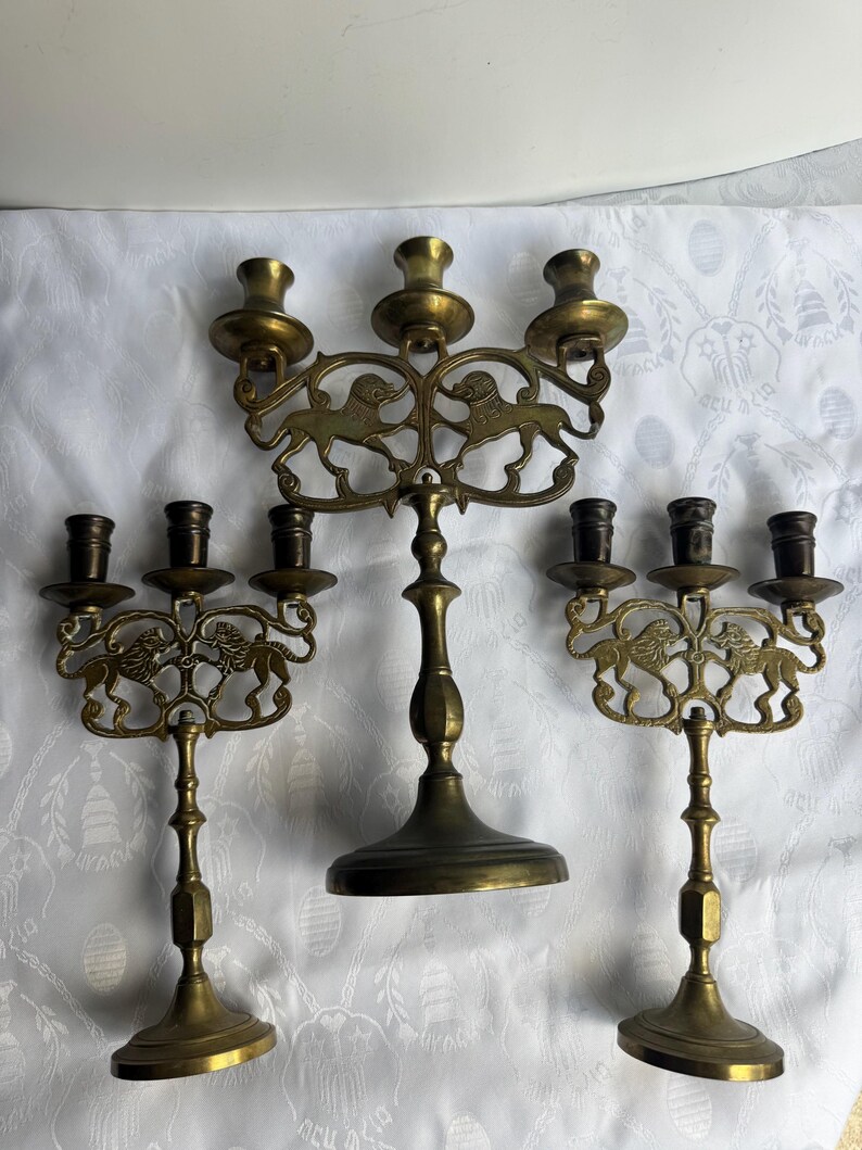 Pu&ograve; includere: Tre candelabri in ottone. Ogni supporto presenta un motivo decorativo a leone e pi&ugrave; portacandele. I candelabri sono di diverse altezze e hanno un'estetica vintage. I candelabri sono su una superficie bianca a motivi.