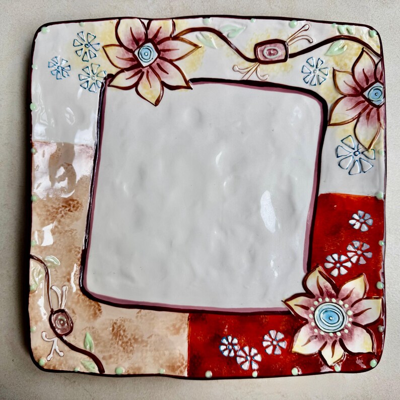May include: A square ceramic plate with a textured white center and a wide border featuring hand-painted floral motifs in shades of red, yellow, and brown. The design includes small blue flower accents and dark brown outlines on a glossy finish.