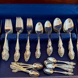 May include: A complete set of ornate silverware, including forks, spoons, and knives, arranged in a blue velvet-lined wooden case. The silverware has a detailed floral pattern on the handles, reflecting light. The set includes various sizes of spoons and forks, and knives.