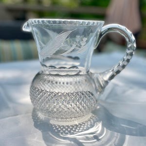 May include: Clear glass creamer with a decorative handle and a diamond-cut base. The creamer features etched feather and floral designs on the upper portion. The handle has a faceted design. A classic piece for serving cream or milk.