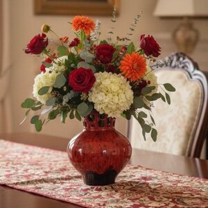 Puede incluir: Un vibrante arreglo floral en un jarr&oacute;n de cer&aacute;mica rojo. El ramo presenta rosas rojas, dalias naranjas e hortensias blancas, con follaje verde. El jarr&oacute;n tiene una superficie texturizada y una base oscura, sobre un camino de mesa estampado.