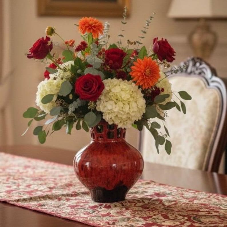 Puede incluir: Un vibrante arreglo floral en un jarr&oacute;n de cer&aacute;mica rojo. El ramo presenta rosas rojas, dalias naranjas e hortensias blancas, con follaje verde. El jarr&oacute;n tiene una superficie texturizada y una base oscura, sobre un camino de mesa estampado.