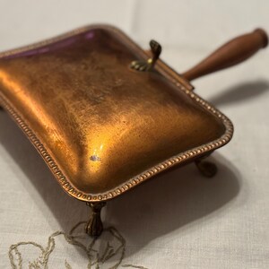 May include: A vintage copper-toned dustpan with a wooden handle and ornate trim. The rectangular pan rests on small, decorative feet. The handle is attached with a brass fitting. The dustpan is designed for collecting dust and debris.