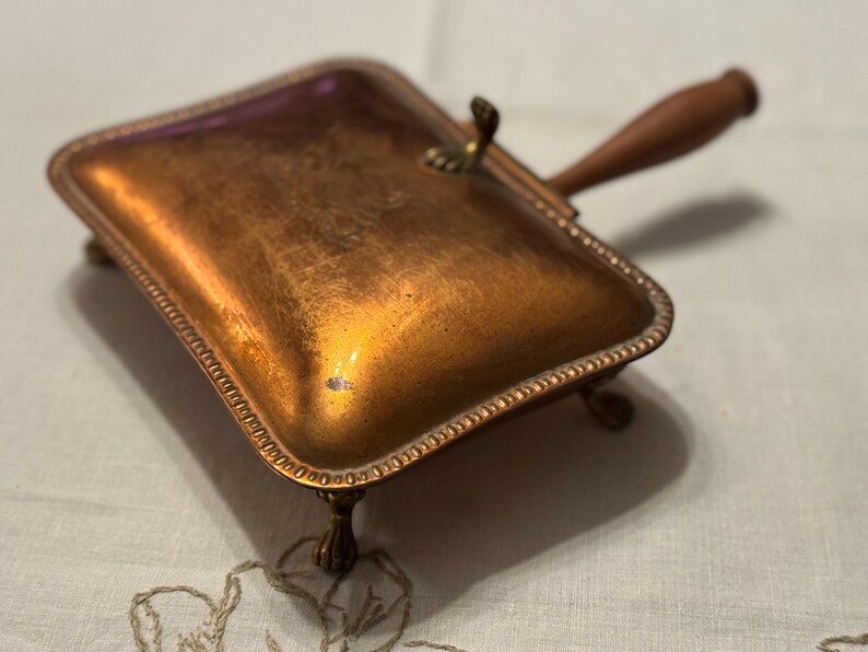 May include: A vintage copper-toned dustpan with a wooden handle and ornate trim. The rectangular pan rests on small, decorative feet. The handle is attached with a brass fitting. The dustpan is designed for collecting dust and debris.