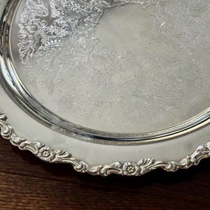 May include: A round silver serving tray featuring an intricate embossed floral and scroll pattern across the center surface. The tray has a scalloped edge with a detailed relief design of small flowers and leaves along the entire rim.