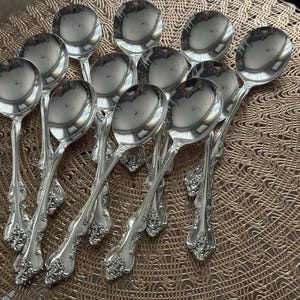 May include: A collection of ten silver soup spoons. Each spoon has a rounded bowl and a decorative handle with floral and scroll detailing. The spoons are arranged on a textured, light brown surface, reflecting the surrounding light.