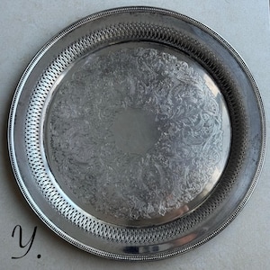 May include: A round silver-toned metal serving tray featuring an intricate engraved floral pattern in the center and a decorative pierced lattice border along the rim. The tray has a smooth, polished finish with a plain circular area in the middle.
