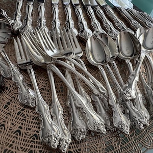 May include: An assortment of antique silverware, including forks, spoons, and knives, arranged on a brown placemat. The silverware has ornate, floral-like detailing and a shiny, silver finish. A soup ladle is also visible.