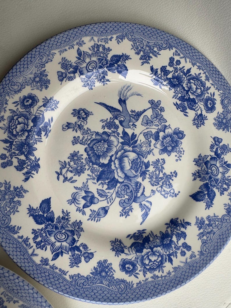 K&ouml;nnte beinhalten: Ein wei&szlig;er Keramik-Dinnerteller mit einem blauen und wei&szlig;en Blumenmuster. Das Muster zeigt einen Vogel und Blumen.