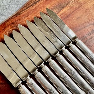 May include: A set of ten silver butter knives with ornate handles. The knives are arranged in a row on a wooden surface.