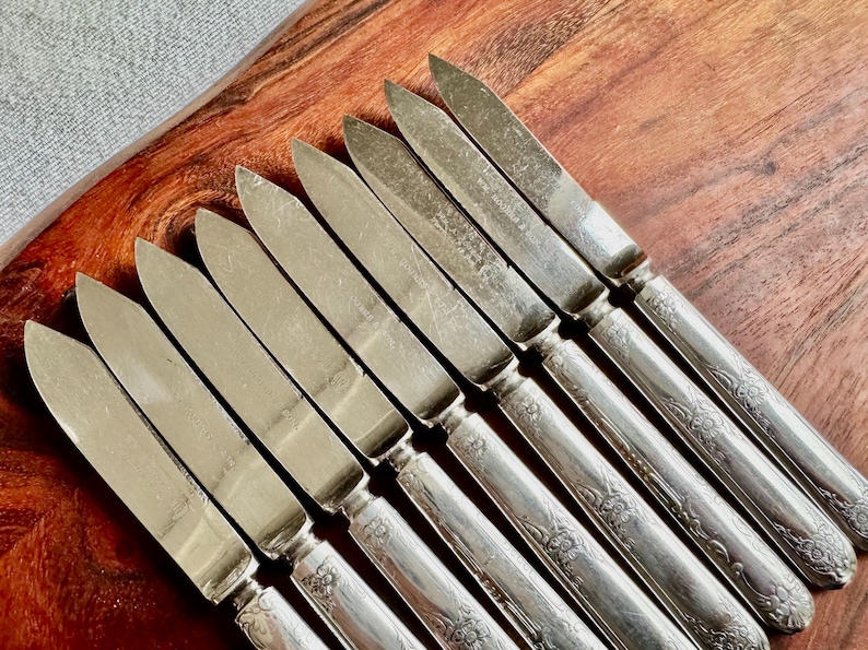 May include: A set of ten silver butter knives with ornate handles. The knives are arranged in a row on a wooden surface.