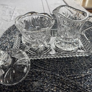 May include: A clear pressed glass creamer, a sugar bowl with a separate lid, and a rectangular tray. Each piece features a faceted starburst pattern and scalloped edges. The set is made of transparent glass with a reflective, textured surface.