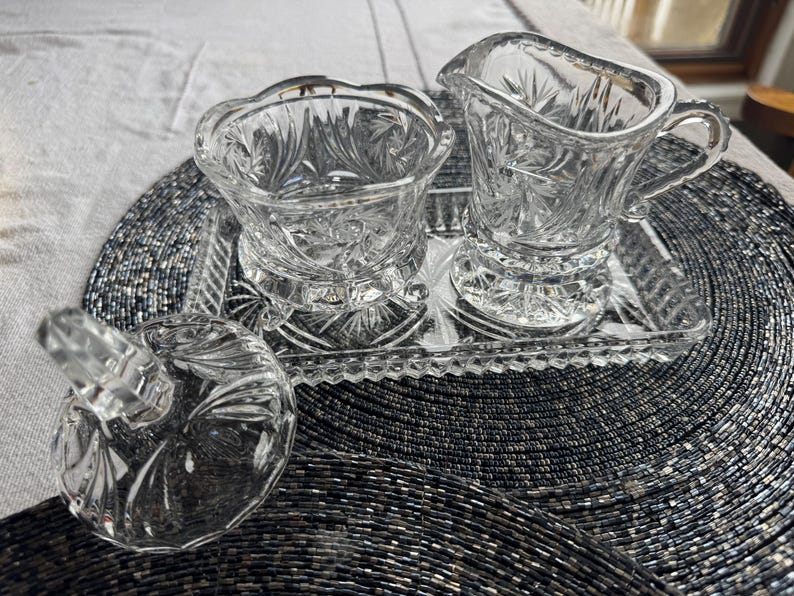 May include: A clear pressed glass creamer, a sugar bowl with a separate lid, and a rectangular tray. Each piece features a faceted starburst pattern and scalloped edges. The set is made of transparent glass with a reflective, textured surface.