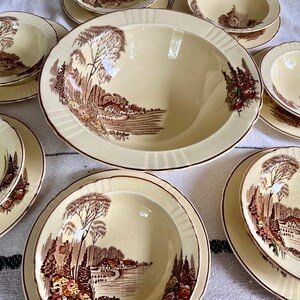 May include: A set of vintage cream-coloured ceramic bowls with brown and green floral patterns. The bowls feature a landscape scene with trees, houses, and a river.
