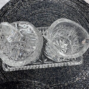 May include: A clear pressed glass creamer and a lidded sugar bowl set resting on a matching rectangular glass tray. Each piece features a detailed starburst and geometric pattern with faceted edges, reflecting light across the transparent surfaces.