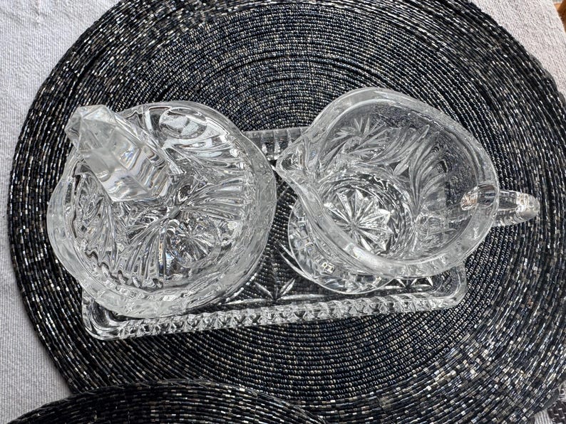 May include: A clear pressed glass creamer and a lidded sugar bowl set resting on a matching rectangular glass tray. Each piece features a detailed starburst and geometric pattern with faceted edges, reflecting light across the transparent surfaces.