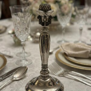May include: A silver-toned metal candlestick holder featuring a flared base with beaded detailing, a slender, vertically ribbed stem, and a decorative top section with a textured, cross-hatched pattern designed to securely hold a single taper candle.