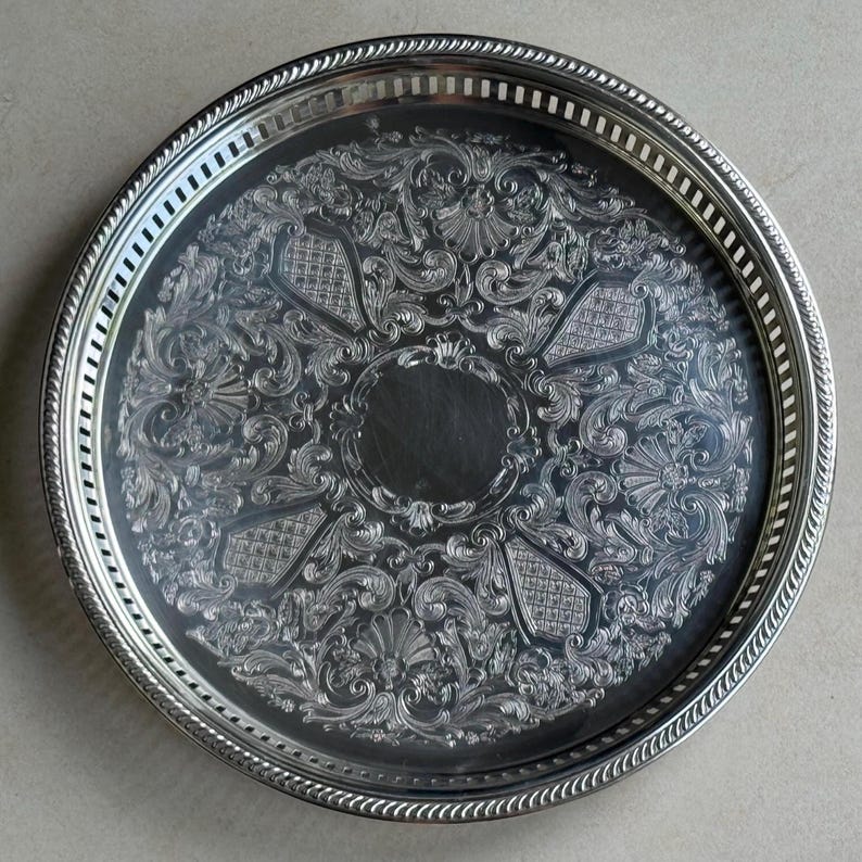 May include: A round silver-toned metal serving tray featuring an intricate embossed floral and scrollwork pattern across the surface. The tray has a raised, pierced gallery rim with a rope-style edge detail surrounding the entire perimeter.