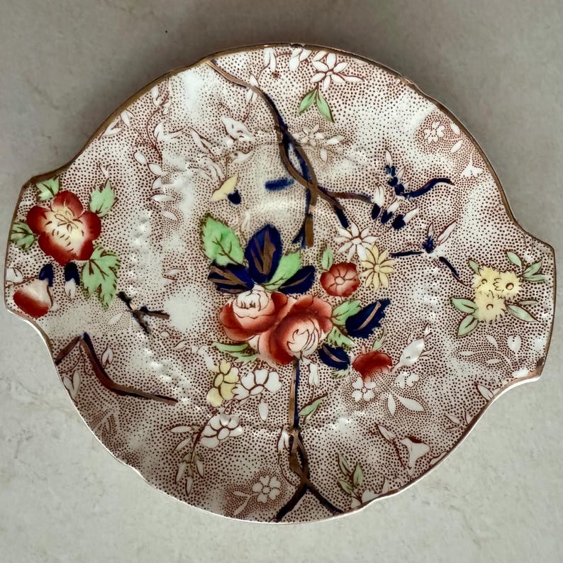May include: A round ceramic serving dish with two side handles, featuring a brown stippled background pattern overlaid with hand-painted floral motifs in red, blue, yellow, and green, accented by dark brown branches and a scalloped edge.
