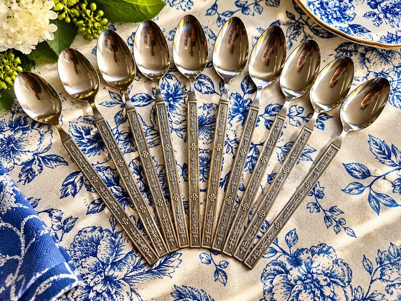 May include: A set of ten stainless steel spoons arranged in a fan pattern. Each spoon features a polished oval bowl and a long, slender handle decorated with an embossed floral and geometric pattern along the entire length of the stem.