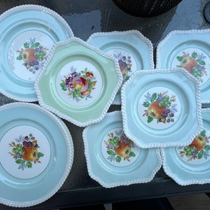 Puede incluir: Un juego de nueve platos de cer&aacute;mica vintage con fondo azul claro y borde blanco. Cada plato presenta un dise&ntilde;o de frutas coloridas con manzanas, uvas y bayas.