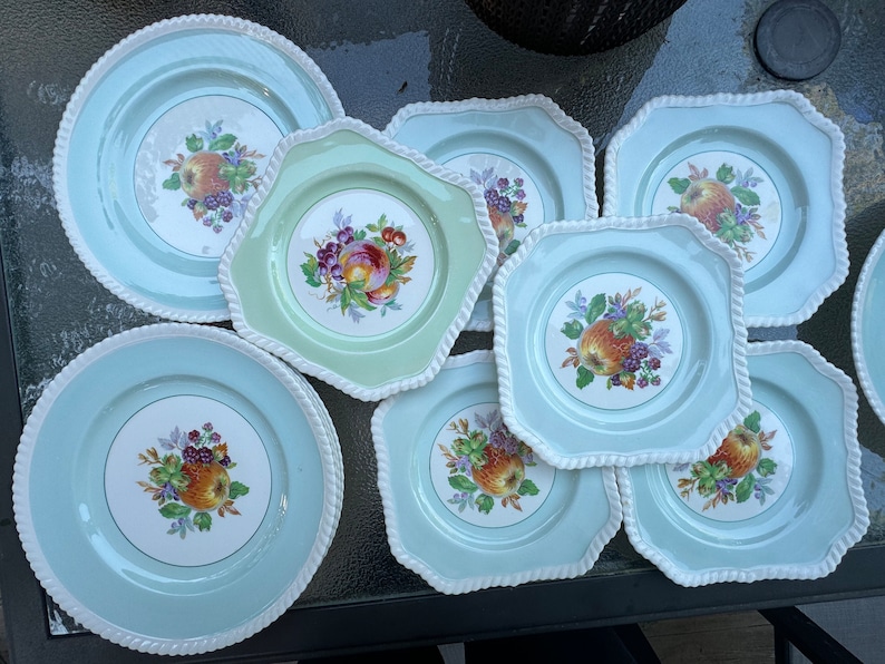 Puede incluir: Un juego de nueve platos de cer&aacute;mica vintage con fondo azul claro y borde blanco. Cada plato presenta un dise&ntilde;o de frutas coloridas con manzanas, uvas y bayas.
