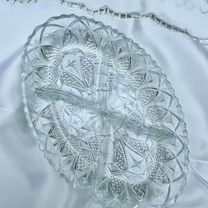 May include: Clear, oval-shaped glass serving dish with three compartments. The dish features a diamond and starburst pattern, with a scalloped edge. The dish is sitting on a white surface.