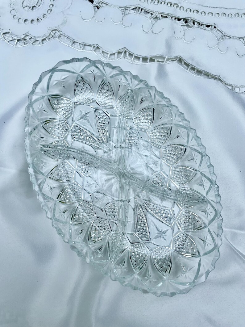 May include: Clear, oval-shaped glass serving dish with three compartments. The dish features a diamond and starburst pattern, with a scalloped edge. The dish is sitting on a white surface.
