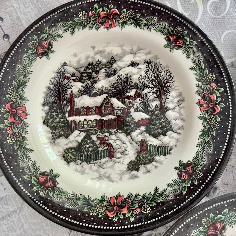 May include: A decorative plate with a winter scene. The plate features a black rim with a white dotted border and a garland of red bows, bells, and greenery. The center depicts a snow-covered village with a house and trees.