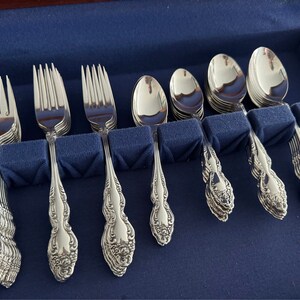 May include: A set of ornate silverware, including forks, spoons, and teaspoons, arranged in a blue velvet-lined case. The silverware has a detailed floral pattern on the handles and a polished, reflective surface. The set is designed for formal dining.