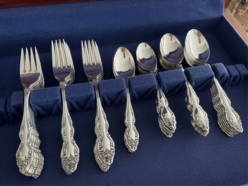 May include: A set of ornate silverware, including forks, spoons, and teaspoons, arranged in a blue velvet-lined case. The silverware has a detailed floral pattern on the handles and a polished, reflective surface. The set is designed for formal dining.