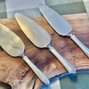 May include: Three silver cake servers with ornate handles rest on a wooden cutting board. The servers have a classic design with a wide, flat blade and a decorative handle. The wood has a natural grain.