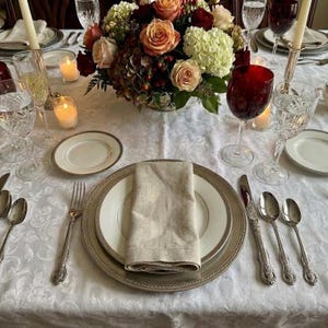 Può includere: Elegante mise en place con un centrotavola floreale. Include piatti, posate, calici e candele. Il centrotavola presenta rose, ortensie e bacche. I piatti hanno un bordo argentato e un tovagliolo di lino piegato.