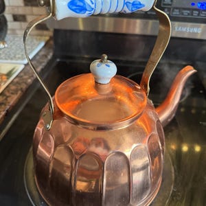 May include: A copper tea kettle with a fluted base, a curved spout, and a metal handle featuring a white ceramic grip with blue floral patterns. The lid is topped with a small, matching white ceramic knob with blue floral accents.