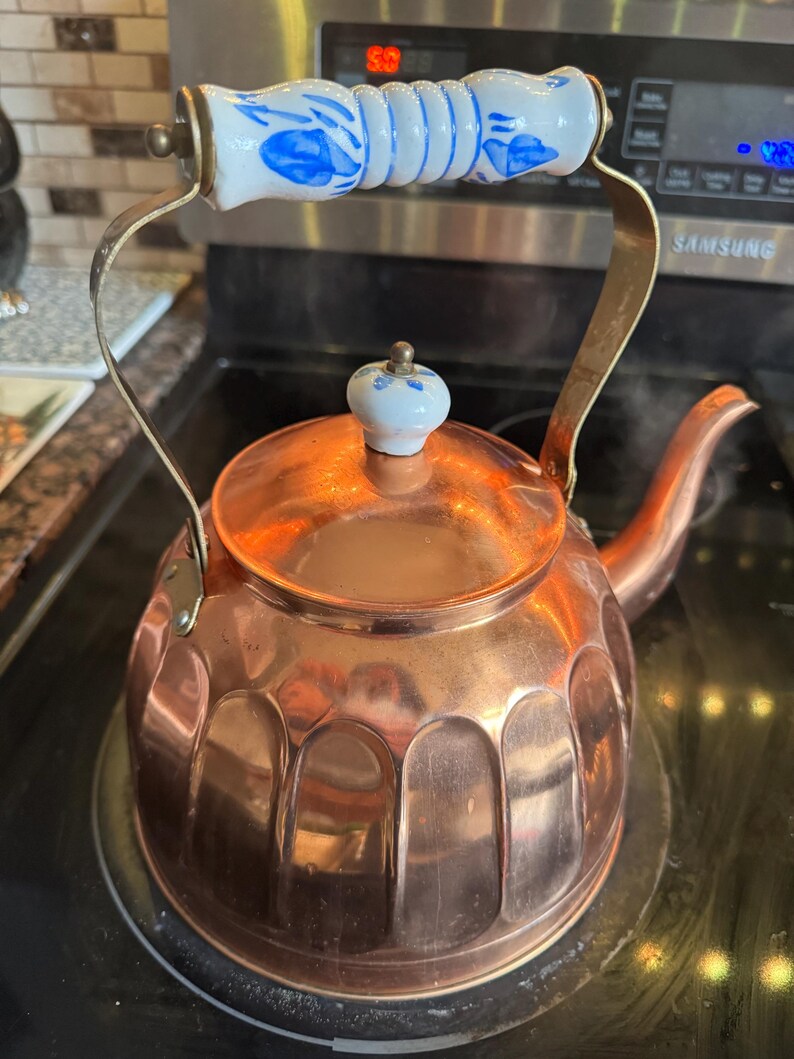 May include: A copper tea kettle with a fluted base, a curved spout, and a metal handle featuring a white ceramic grip with blue floral patterns. The lid is topped with a small, matching white ceramic knob with blue floral accents.