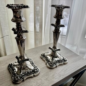 May include: A pair of silver-toned metal candlesticks featuring ornate, embossed floral detailing on the square bases and stems. Each candlestick has a flared top rim and a tapered central column, showcasing a polished, reflective metallic finish.