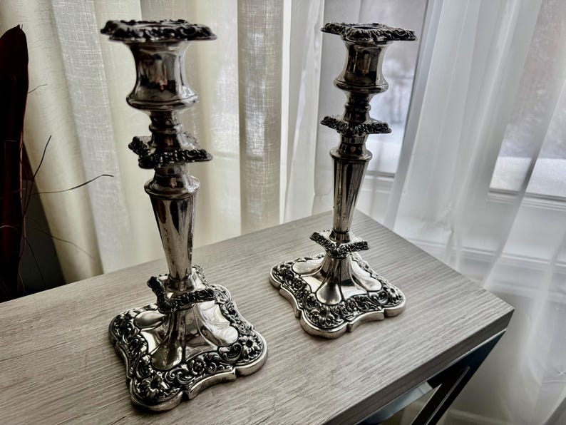 May include: A pair of silver-toned metal candlesticks featuring ornate, embossed floral detailing on the square bases and stems. Each candlestick has a flared top rim and a tapered central column, showcasing a polished, reflective metallic finish.