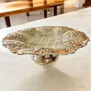 May include: Silver-plated serving dish with a scalloped edge and floral relief design. The dish is raised on a round pedestal base. The interior has a rippled texture. The dish is displayed on a white surface.