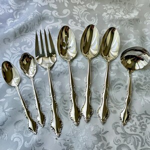 May include: A collection of silver-colored flatware, including spoons, a fork, and a ladle, arranged on a patterned, light-colored surface. The silverware has ornate handles and reflective surfaces, suggesting a formal dining setting.