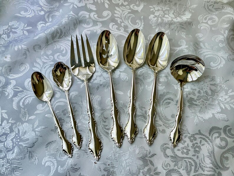 May include: A collection of silver-colored flatware, including spoons, a fork, and a ladle, arranged on a patterned, light-colored surface. The silverware has ornate handles and reflective surfaces, suggesting a formal dining setting.