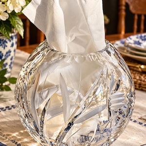 May include: A spherical clear crystal tissue holder featuring intricate etched leaf and floral patterns on its exterior surface. The container is filled with a white paper tissue that extends from the circular opening at the top.